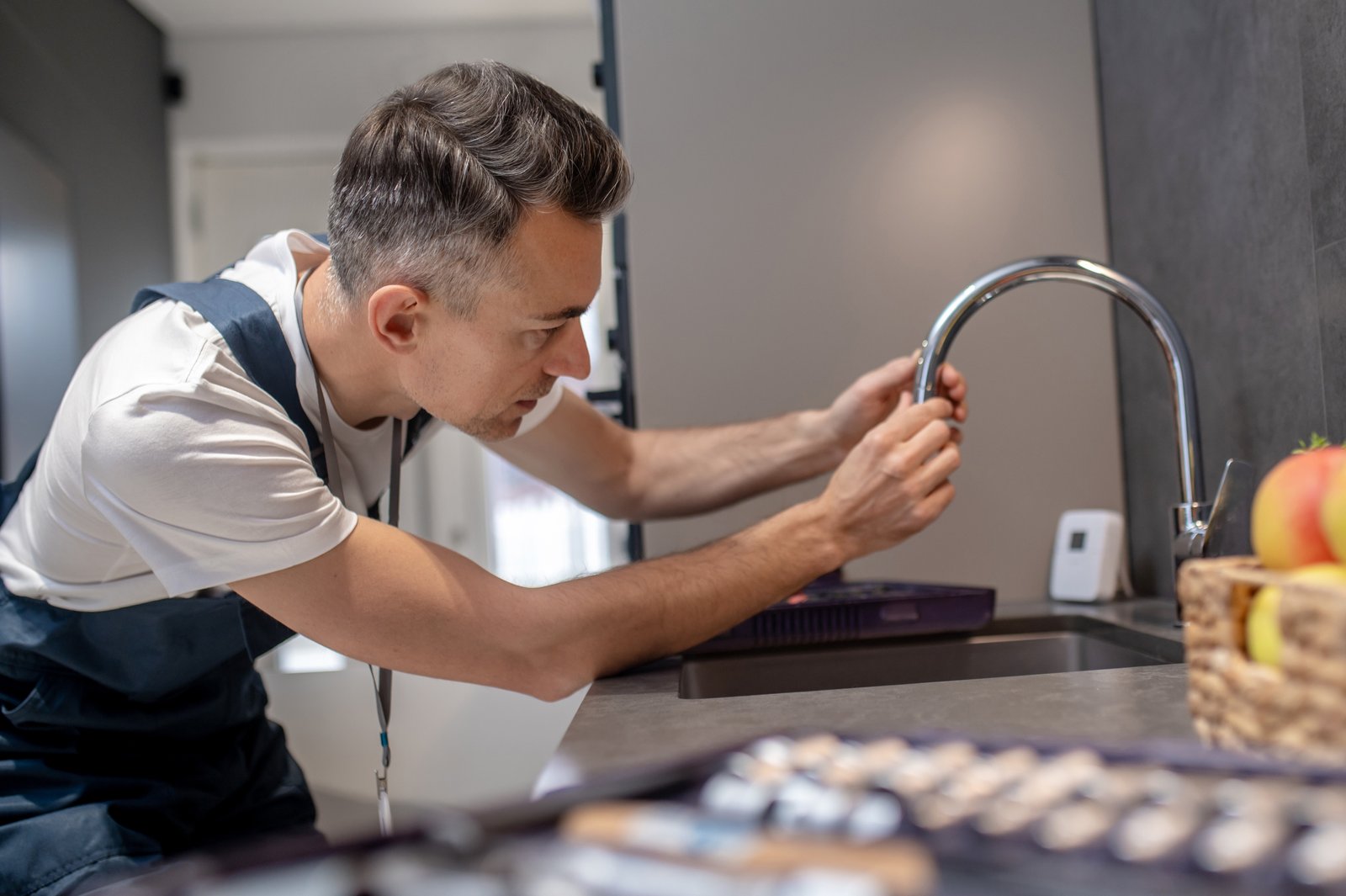 Plumber in an apron carefully installing or repairing a curved kitchen faucet over a sink in a modern kitchen, with tools in the foreground and a focused, professional atmosphere